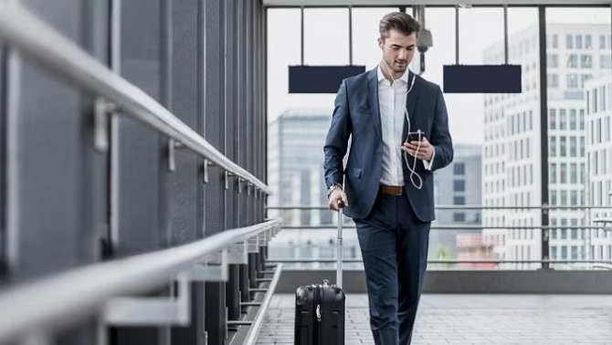 Man standing next to suitcase while looking at a smartphone in an airport representing the importance of a  business travel checklist for a seasoned pro or first-time traveller alike.
