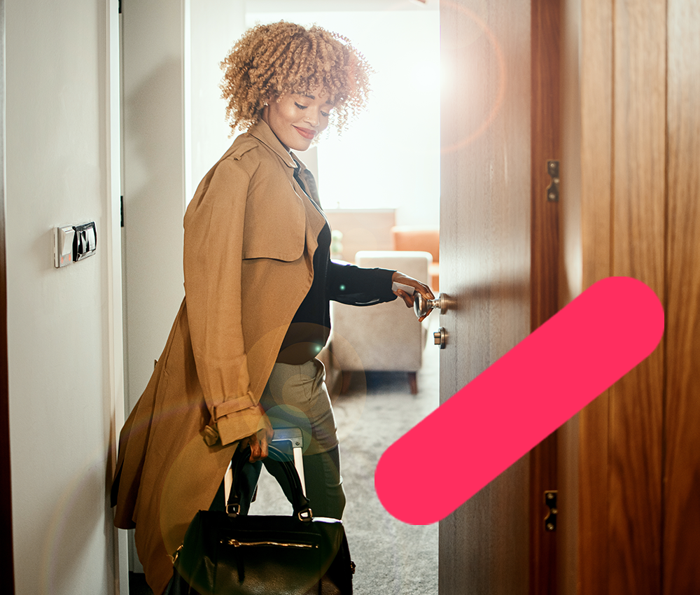 Smiling young woman with curly hair in trench coat opens hotel door carrying suitcase in sunlight