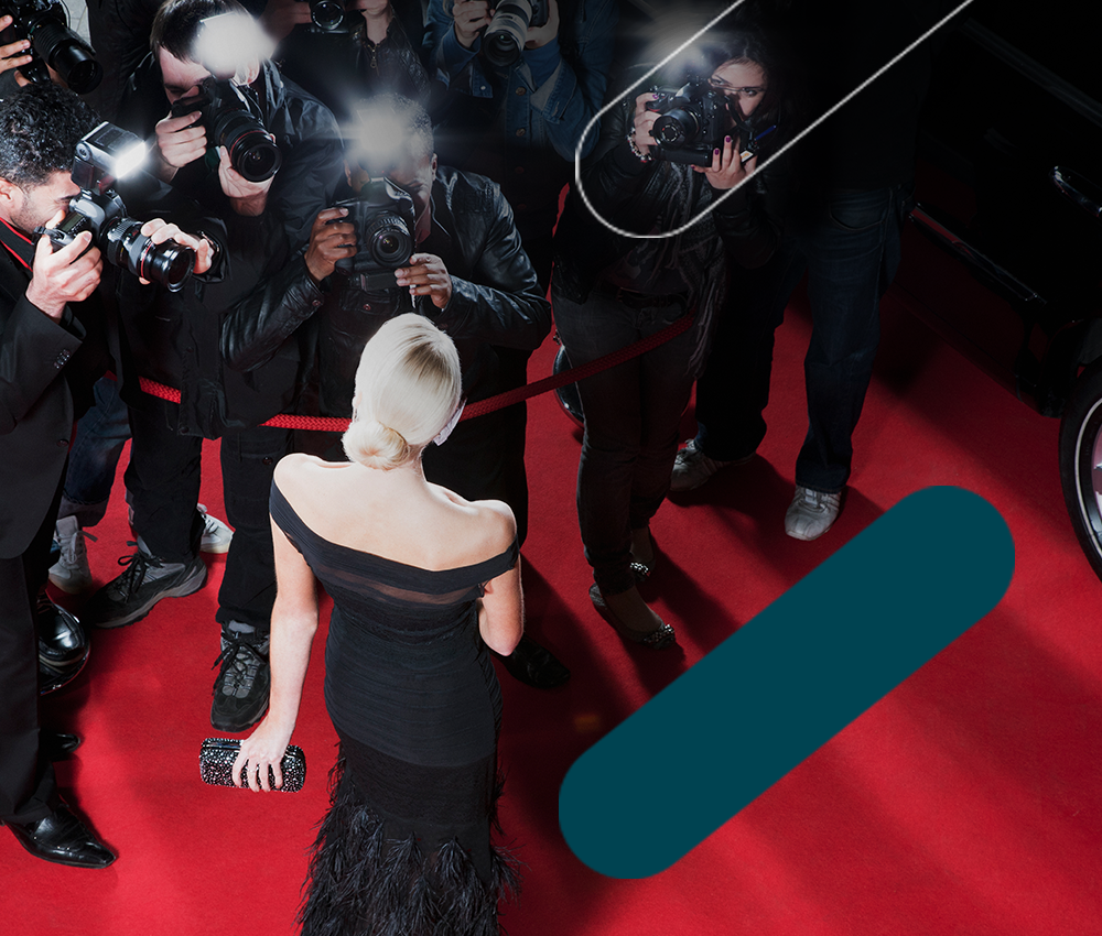 Back view of blonde celebrity in black gown on red carpet facing flashing paparazzi cameras