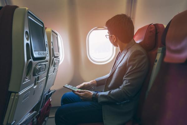 Business traveler in suit seated by airplane window holding tablet, gazing outside.